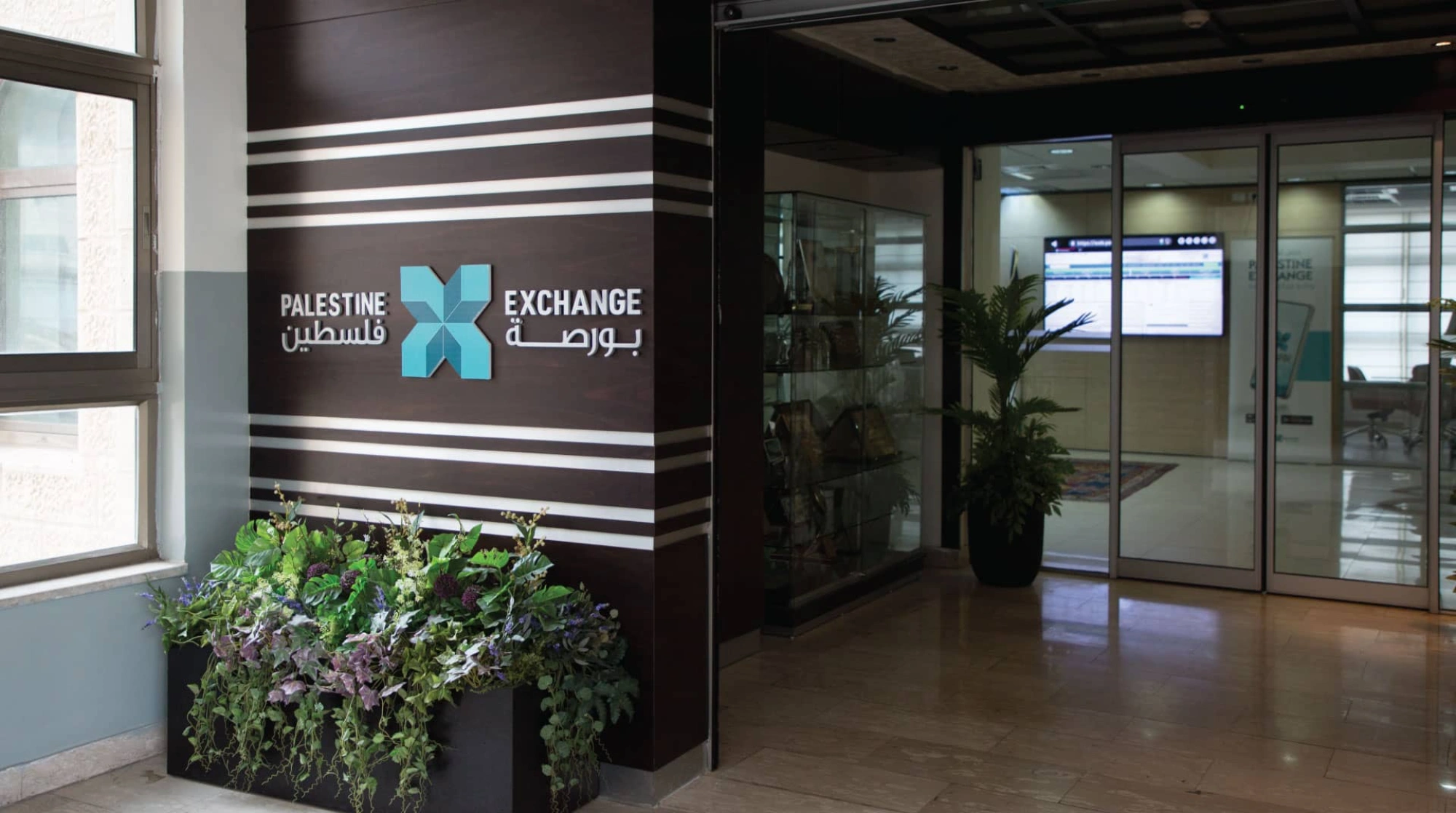 The entrance hall of the Palestine Exchange: its name and star mark mounted in Arabic and English on a dark wood wall beside the glass trading-floor doors.